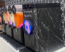 Load image into Gallery viewer, A close-up of a black marble-patterned beverage dispenser with a circular blue-lit button, next to other drink dispensers containing orange and pink drinks. Handwritten text on the front reads 'Strawberry Margarita'.