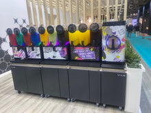 Load image into Gallery viewer, A freestanding multi-drink slushie machine display at an indoor trade show. The unit has multiple colorful drink reservoirs with black dispensing heads, a large front display panel with graffiti-style graphics and a circular dial, and dark gray wheeled cabinets underneath.
